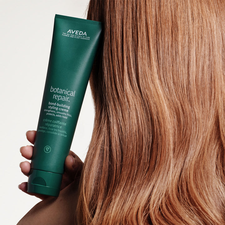 image of a hand holding a tube of aveda botanical repair bond-building styling creme against smooth, auburn hair.